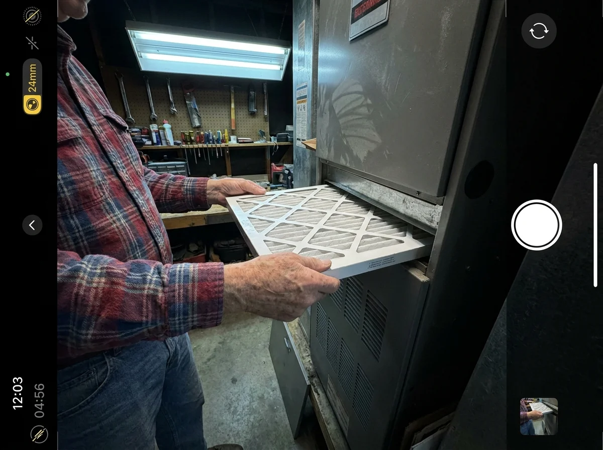 A close-up shot of a senior person's hands sliding a standard air filter into a home furnace unit in a garage.