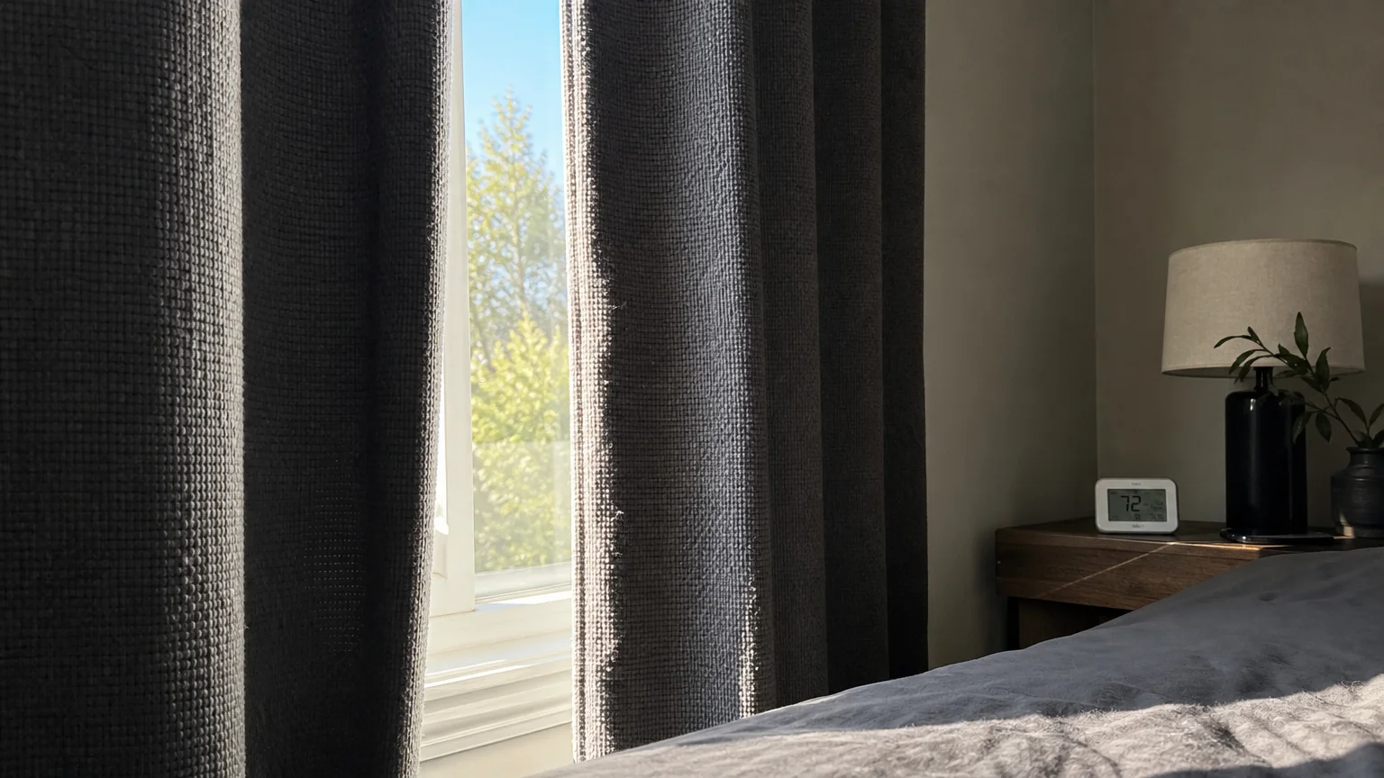 A close-up of thick grey thermal curtains partially drawn to block bright sunlight in a home bedroom.