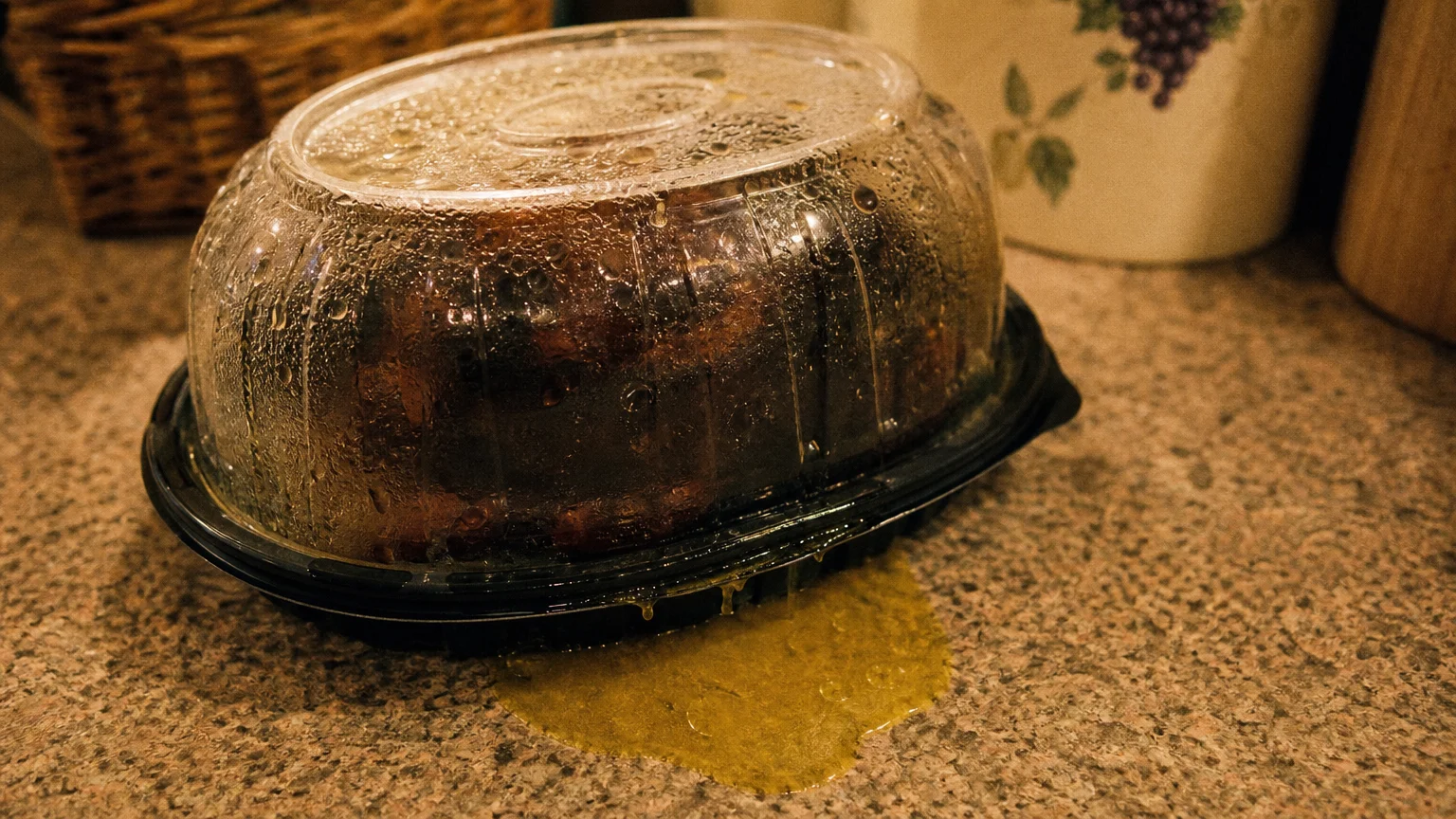 A close-up photo of a greasy plastic rotisserie chicken container with yellow grease leaking onto a laminate counter.