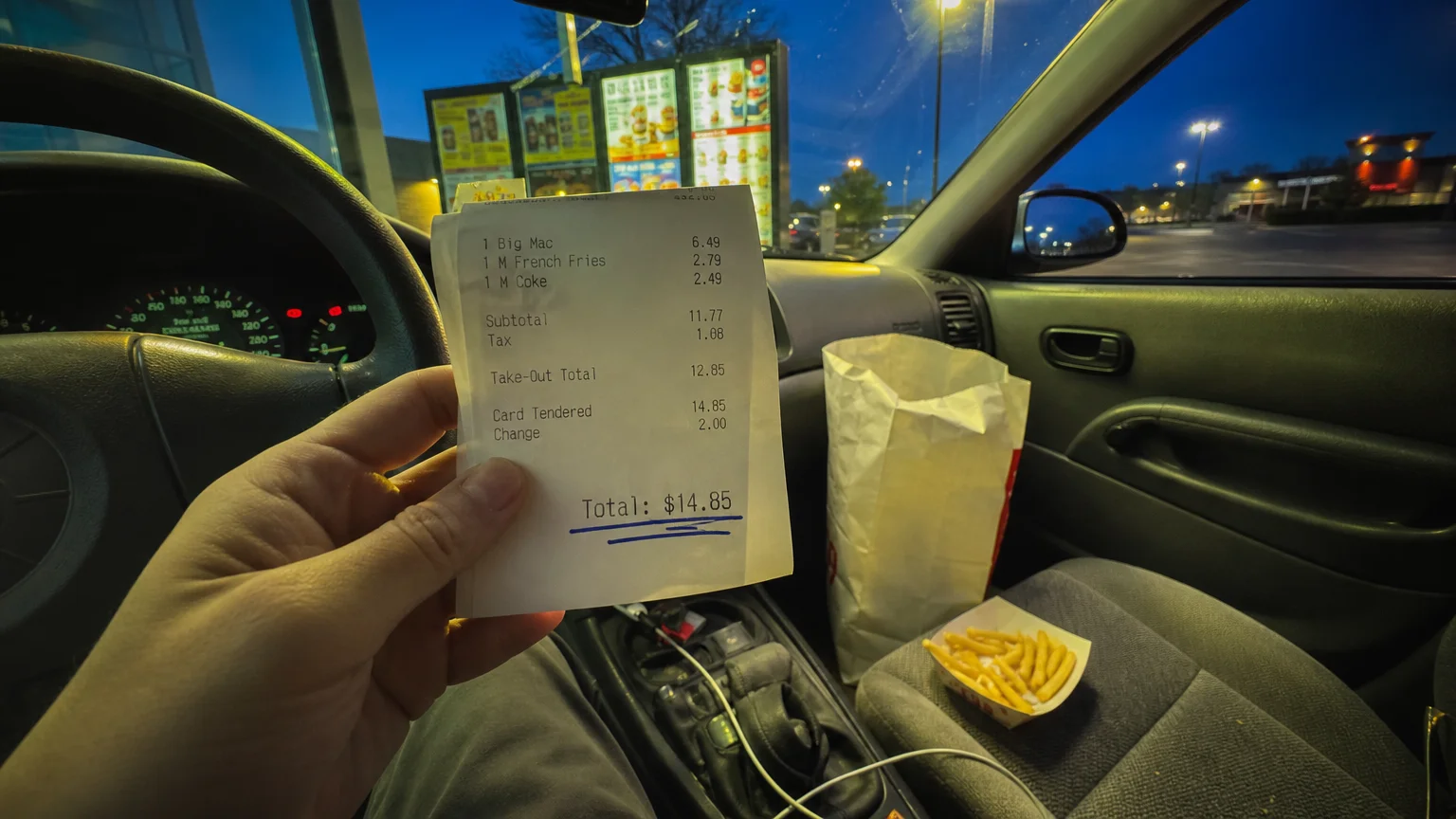 A first-person view of a fast food receipt for $14.85 inside a car at night, with a plain food bag on the seat.
