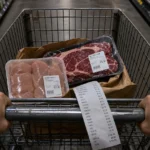 A grocery cart containing expensive packages of meat and a long receipt, shot from a first-person perspective in a grocery store.