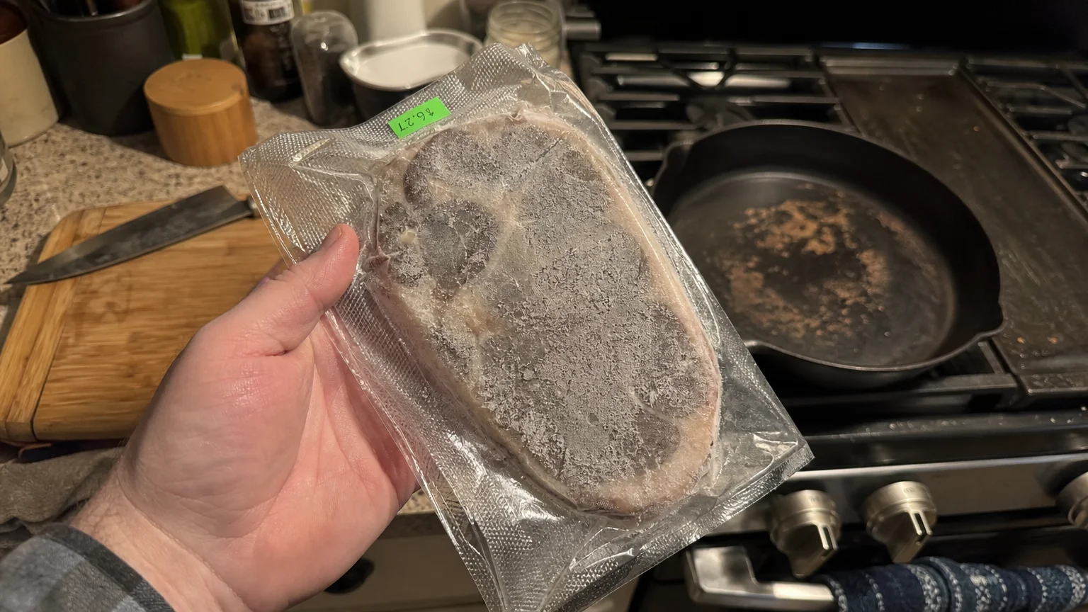 A hand holding a thin, gray, frozen steak in vacuum packaging with visible ice crystals in a home kitchen.