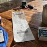 A Walmart receipt and several store-brand products like spices and medicine sit on a sunlit wooden kitchen table next to a coffee mug.