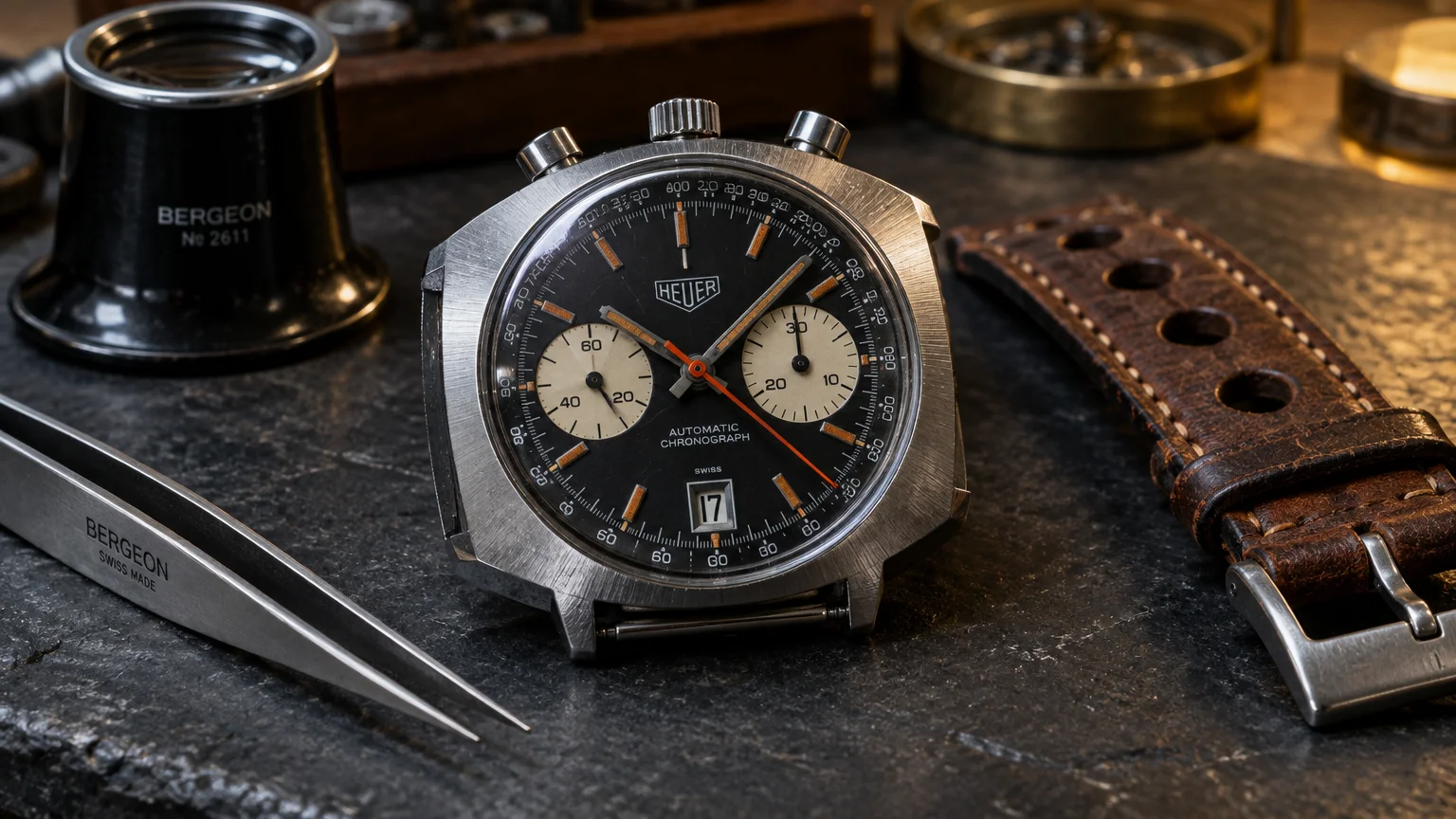 Vintage Heuer chronograph watch head on a workbench beside watchmaker tweezers, a loupe, and a brown leather strap.